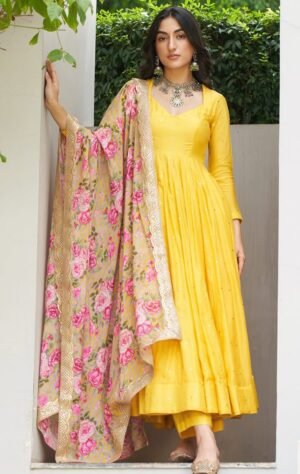 long-sleeved yellow Anarkali dress with a flared skirt and a V-neckline. The dress has subtle, scattered embellishments. She holds a contrasting dupatta with a bold pink floral pattern