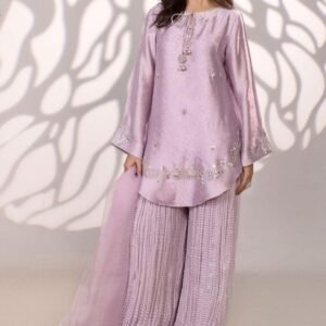 She is modeling a three-piece lavender silk suit. The short, flared kurti has long, wide sleeves and is embellished with delicate silver embroidery and sequins around the neckline and hem. It is paired with wide-legged, heavily crinkled palazzo pants and a sheer lavender dupatta.