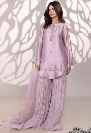 She is modeling a three-piece lavender silk suit. The short, flared kurti has long, wide sleeves and is embellished with delicate silver embroidery and sequins around the neckline and hem. It is paired with wide-legged, heavily crinkled palazzo pants and a sheer lavender dupatta.