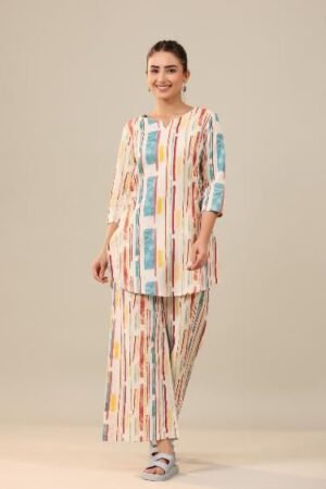 A smiling woman with her hair in a bun is wearing a casual two-piece loungewear set. The set is made of off-white khadi cotton and features a vibrant, abstract vertical stripe print in colors like red, yellow, and blue. The top is a short tunic with a V-neck and three-quarter sleeves, and the bottom is a pair of wide-leg palazzo pants. She is standing against a neutral-colored background.
