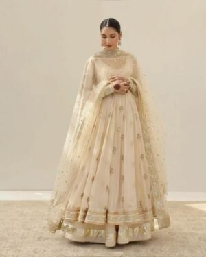 A full-length photo of a woman wearing a cream-colored Anarkali suit with intricate gold embroidery. The dress has a long, flowing skirt and is paired with a matching dupatta adorned with small gold embellishments.