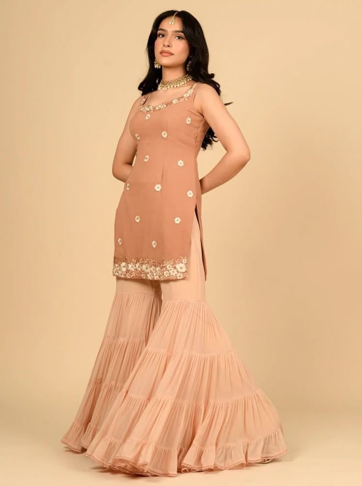 A South Asian woman models a peach-colored sharara suit. The sleeveless kurta is adorned with delicate white floral embroidery on the bodice and hem. The tiered sharara pants are a slightly lighter shade of peach.